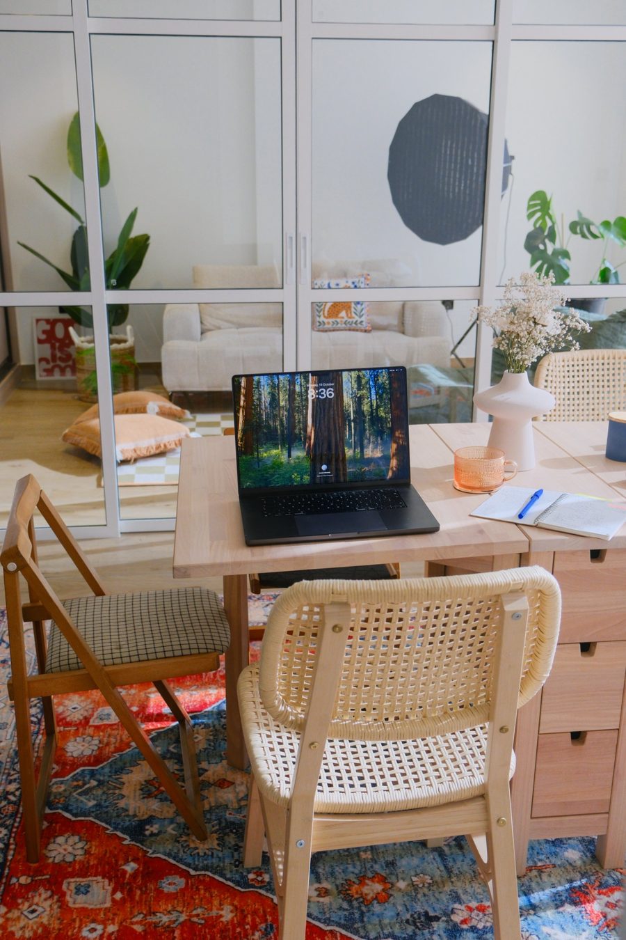 A laptop, notebook and vase of dried flowers on the coworking table, with the glass-walled studio behind